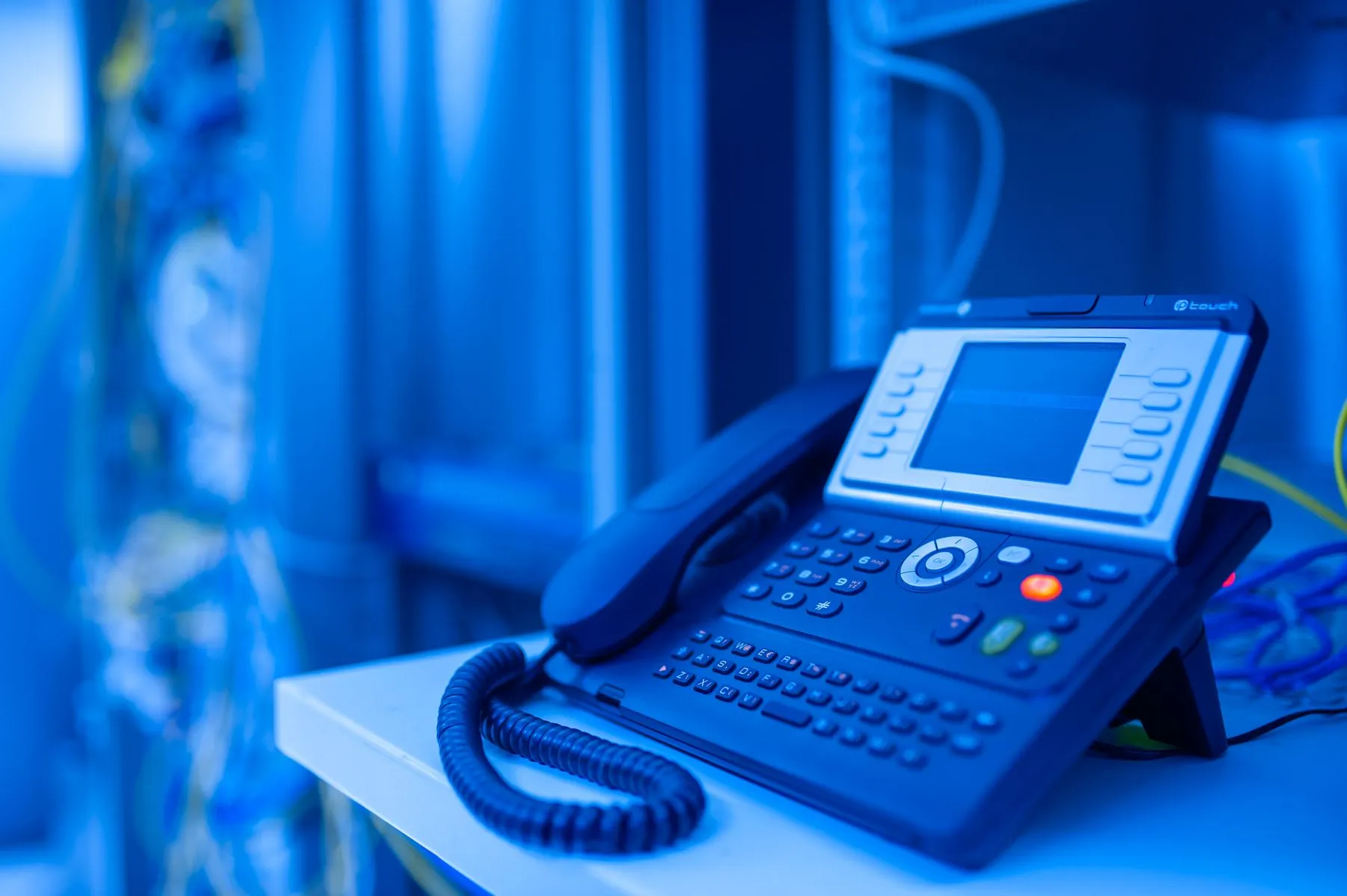 IP desk phone with a display screen and handset on a desk, lit blue