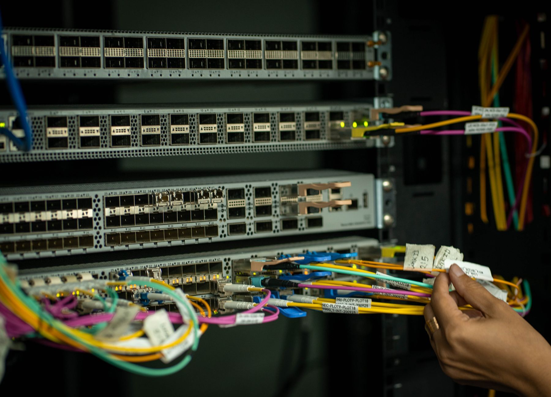 Hand holding labeled fiber patch cords at a network switch in an equipment rack