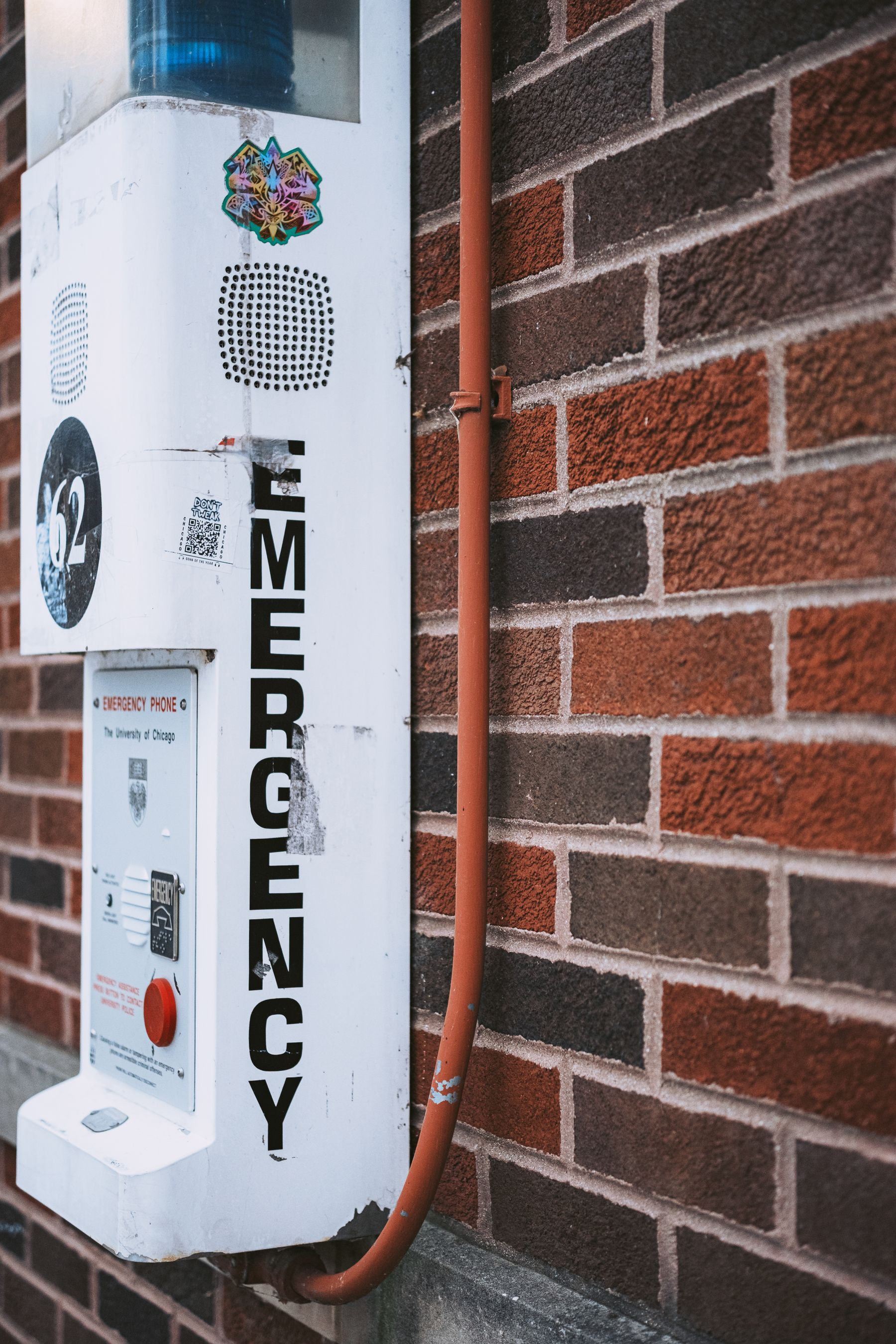 White emergency call station column with a blue beacon and vertical EMERGENCY lettering, mounted on a brick wall